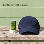 SPF 50 mineral sun stick on a weathered wooden bench beside a navy baseball cap. For the seventh inning.