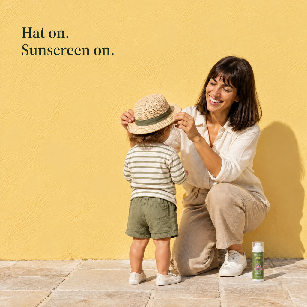 A woman kneels to set a straw sun hat on a toddler's head, the baby sunscreen pump on the ground beside her. Hat on. Sunscreen on.