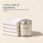Jar of baby tallow balm beside a stack of folded white cloths. A short stack of ingredients. Grass-fed tallow, zinc oxide, calendula, shea butter, jojoba oil, beeswax.