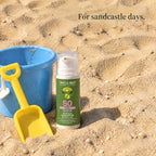 Baby mineral sunscreen pump bottle in the sand beside a blue bucket and yellow spade. For sandcastle days.