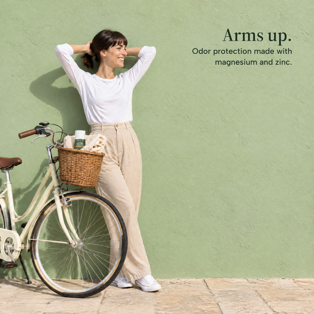 A woman stretches with her hands behind her head beside a bicycle, the deodorant in its basket. Arms up. Odor protection made with magnesium and zinc.