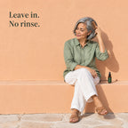 A woman with silver hair combs it while sitting on a ledge, the spray bottle beside her. Leave in. No rinse.