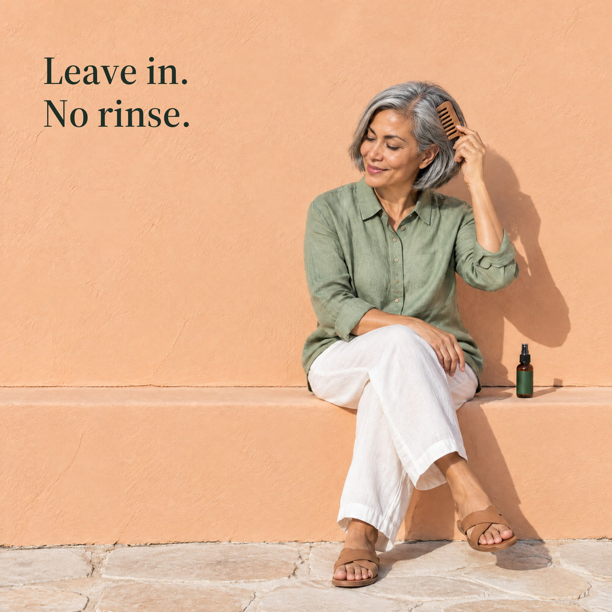 A woman with silver hair combs it while sitting on a ledge, the spray bottle beside her. Leave in. No rinse.