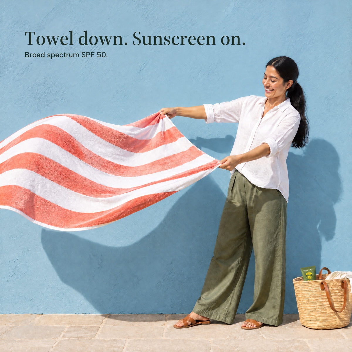 A woman shakes out a red and white striped beach towel by a blue wall, the sunscreen tube in a basket at her feet. Towel down. Sunscreen on. Broad spectrum SPF 50.