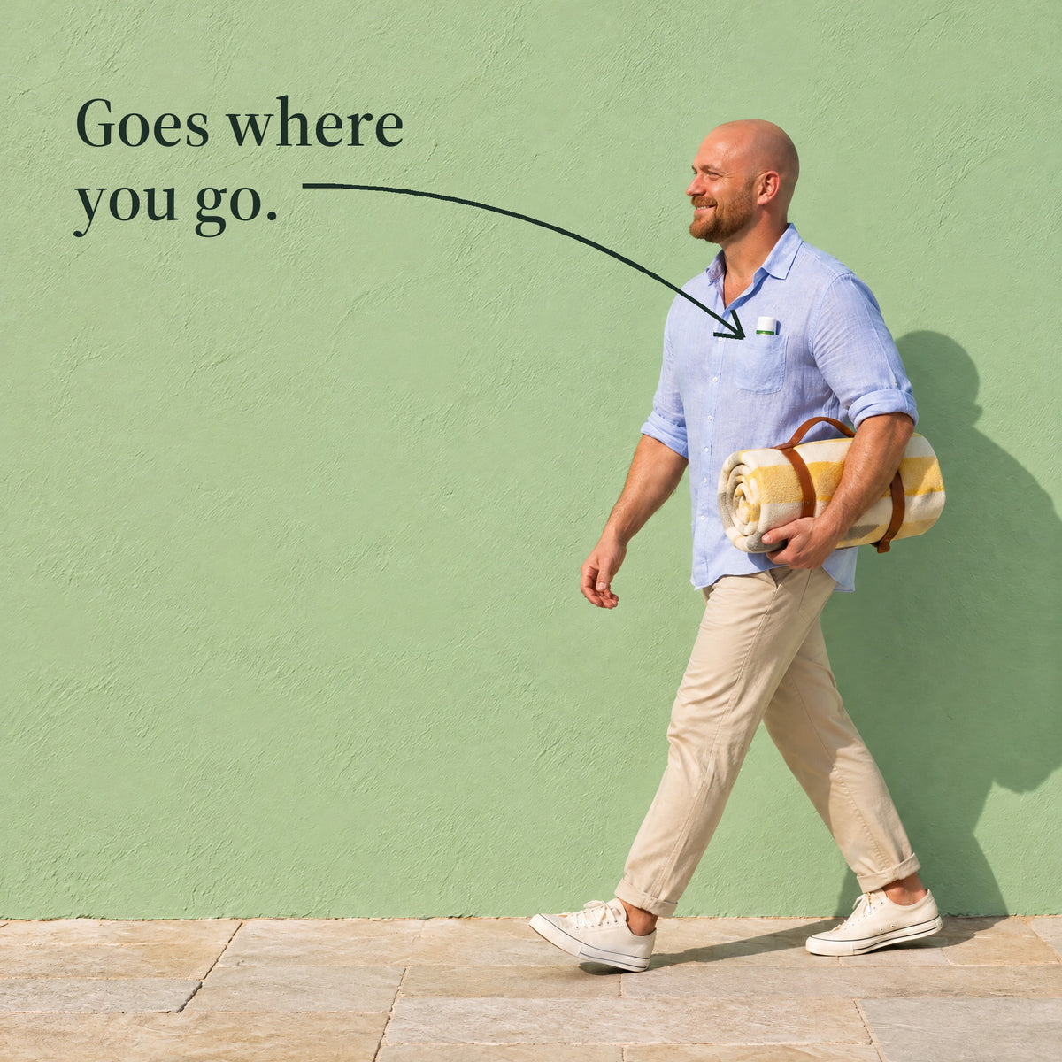 A man walks past a green wall with a rolled picnic blanket, the sun stick in his shirt pocket. Goes where you go.