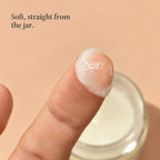 A fingertip holding a small scoop of baby tallow balm above the open jar. Soft, straight from the jar.