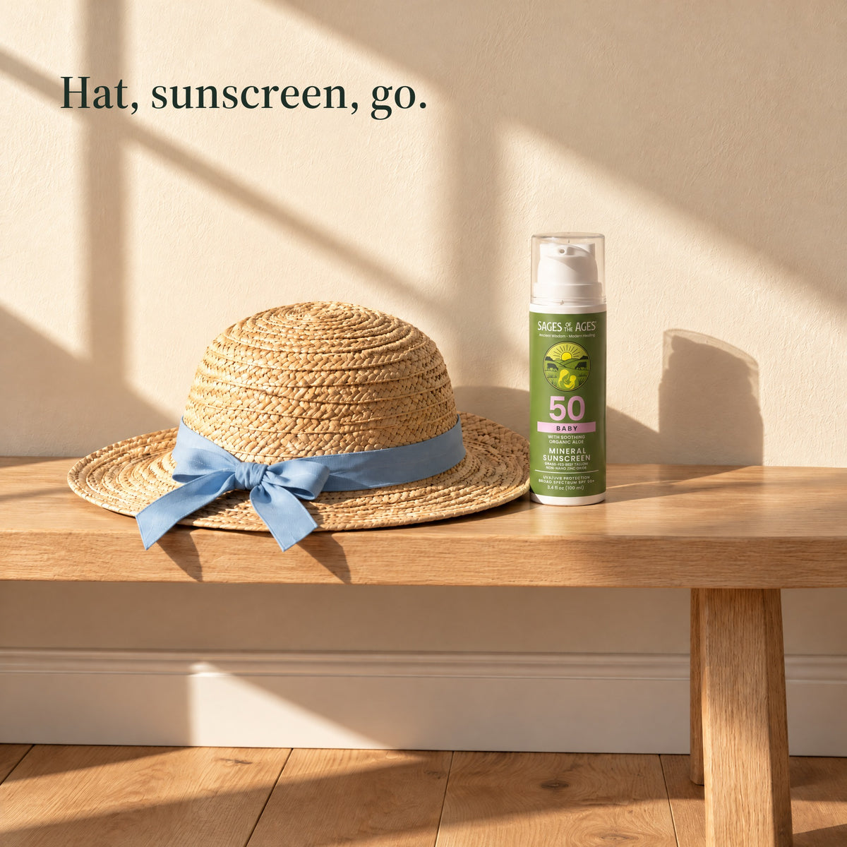 Baby mineral sunscreen pump bottle on a wooden bench next to a straw hat with a blue ribbon. Hat, sunscreen, go.