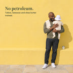 A father holds his baby at his shoulder by a yellow wall, the jar of balm on the ledge beside them. No petroleum. Tallow, beeswax and shea butter instead.