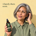 A woman with grey hair holds up the bottle of rice water hair spray against a yellow wall. A barely-there scent.