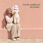 A mother in a pink headscarf sits on a bench lifting her baby, the jar of balm beside her. For the smallest in the family.