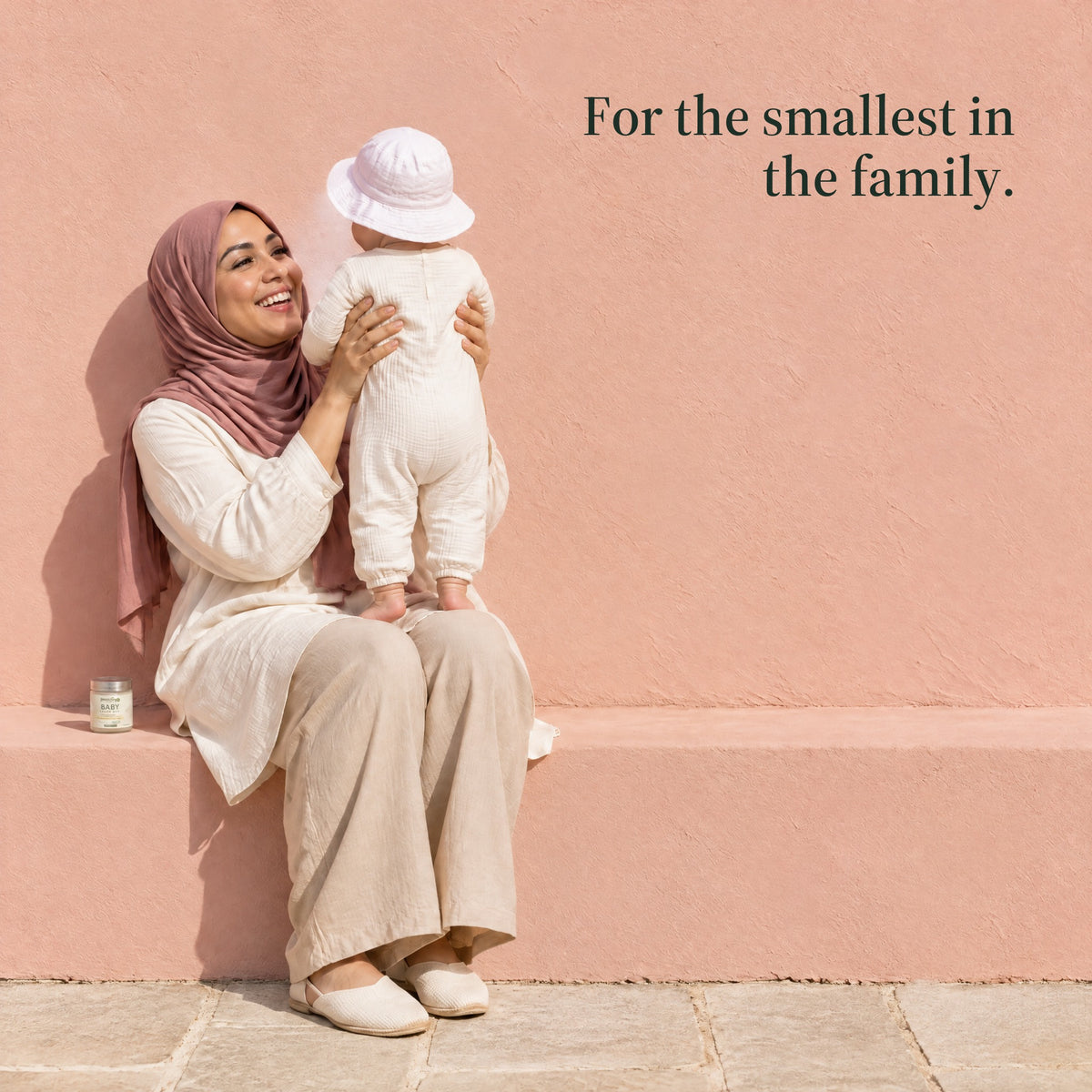 A mother in a pink headscarf sits on a bench lifting her baby, the jar of balm beside her. For the smallest in the family.