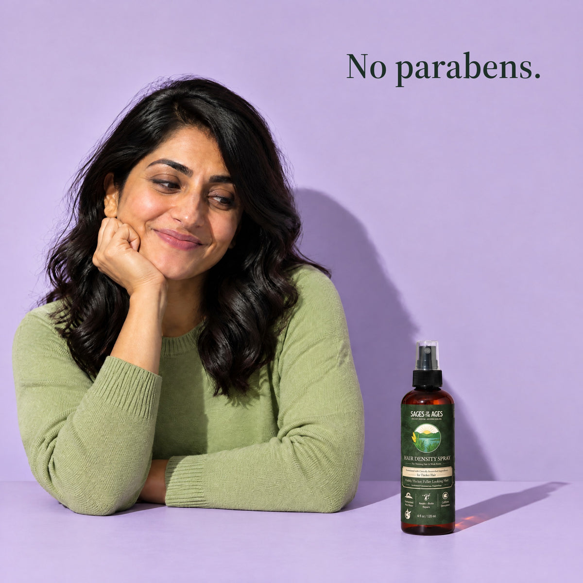 A woman with dark wavy hair rests her chin on her hand, the spray bottle beside her on a lilac background. No parabens.