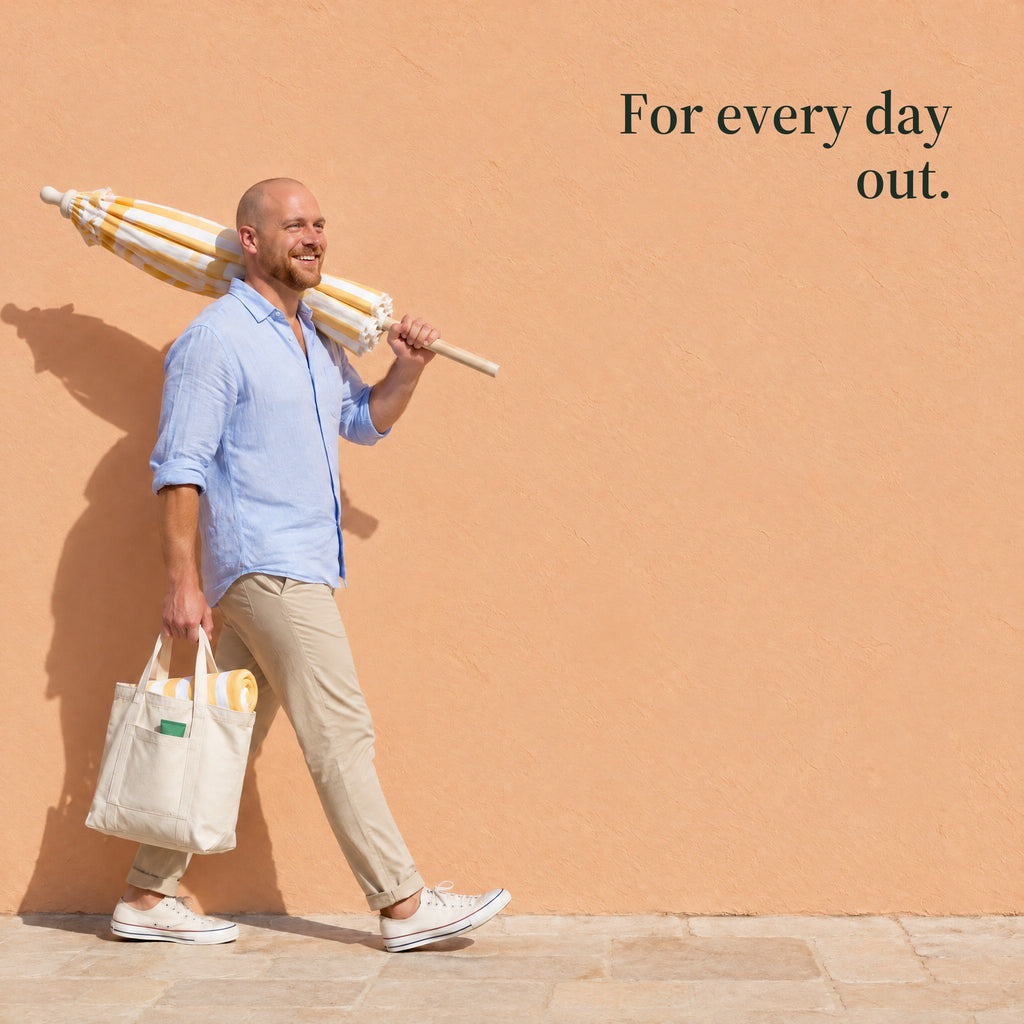 A man walks past a peach wall with a beach umbrella on his shoulder and a canvas tote in his hand. For every day out.