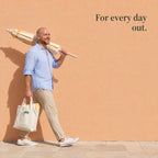 A man walks past a peach wall with a beach umbrella on his shoulder and a canvas tote in his hand. For every day out.