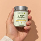 A hand holds the jar of baby tallow balm against a peach wall.
