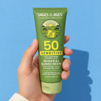 A hand holds up the tube of SPF 50 mineral sunscreen lotion against a blue wall.